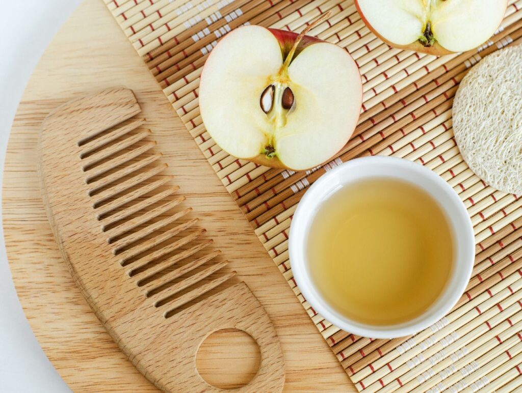 Ein Apfel, Apfelessig und ein Holzkamm liegen auf einer Holzplatte.