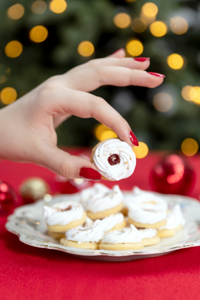 Frauenhand hält einen Weihnachtskeks zwischen Daumen und Zeigefinger, dahinter Vulkanlandkrapferln auf Teller